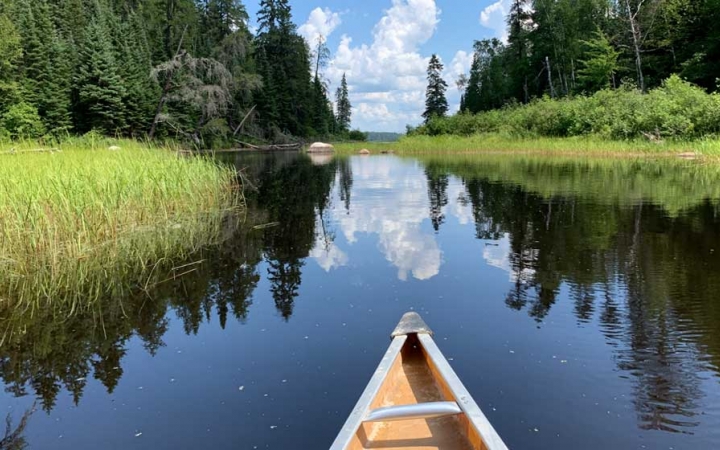 In the foreground, the tip of a canoe appears above very calm water, reflecting the grasses and trees on the shore, and the blue sky and clouds above. 